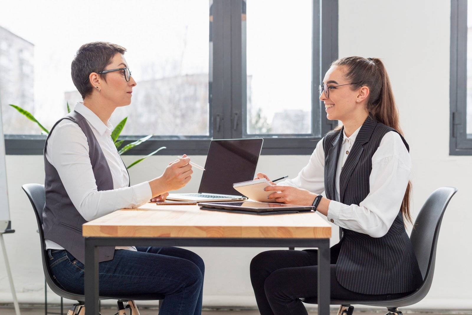 Two women having a discussion or interview in an office setting, sitting at a table with a laptop and notepad.