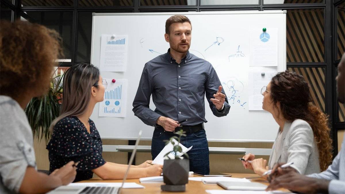 Man giving a presentation to a group of women seated at a table, with charts and notes on a whiteboard behind him.